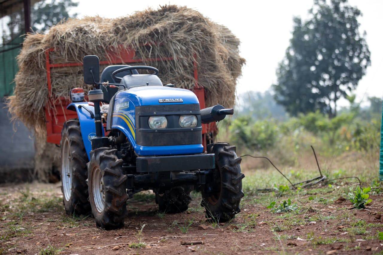 A blue tractor loaded with hay bales in a rural setting, showcasing agriculture and farming.
