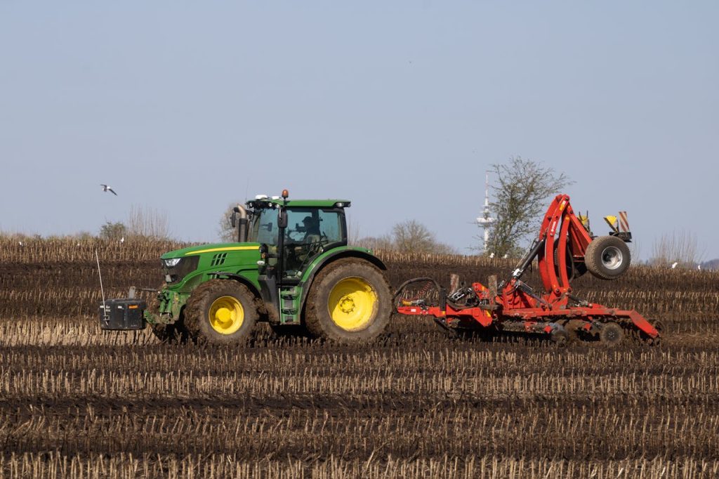 A green tractor with a red harrow plows a brown farmland under clear blue sky.
