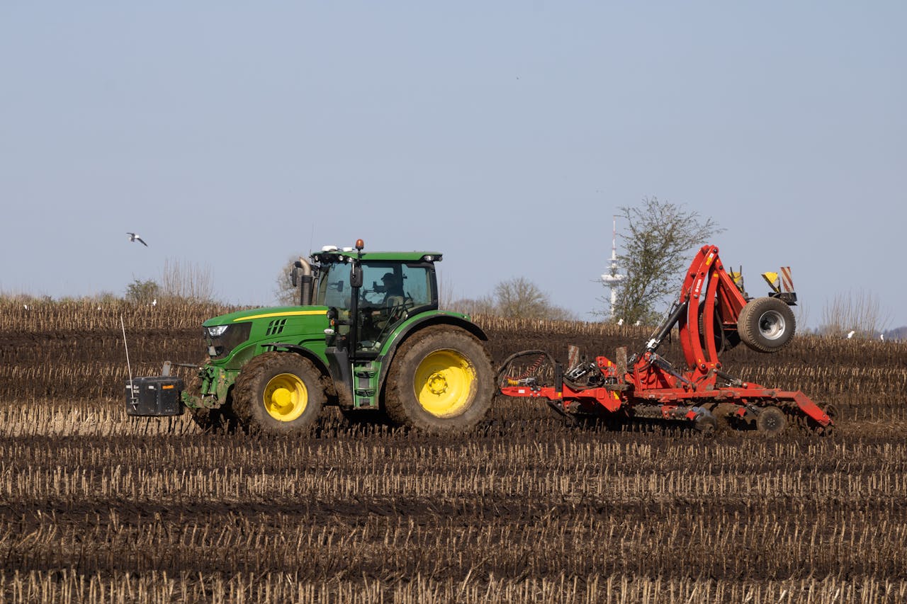 A green tractor with a red harrow plows a brown farmland under clear blue sky.